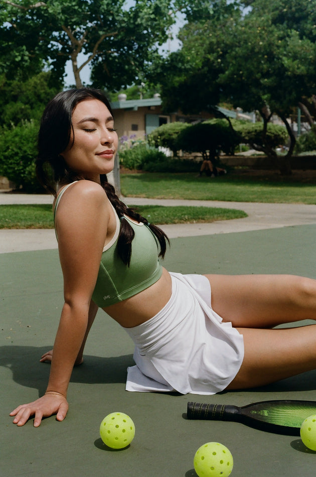 Woman in green sports bra and white shorts sitting on a tennis court with pickleballs around her.