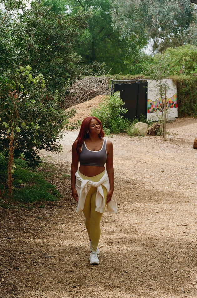 Woman walking on a dirt path in a natural setting with trees and a colorful wall in the background.